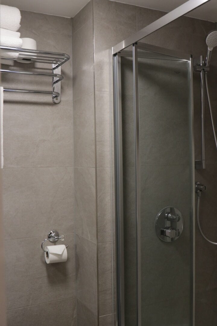 A modern bathroom with gray tiled walls, a glass shower enclosure, a metal towel rack with white towels, and a toilet paper holder mounted on the wall.