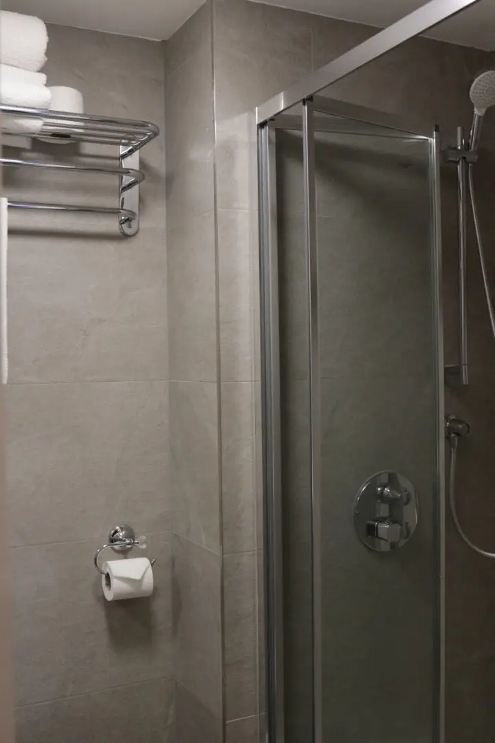 A modern bathroom with gray tiled walls, a glass shower enclosure, a metal towel rack with white towels, and a toilet paper holder mounted on the wall.