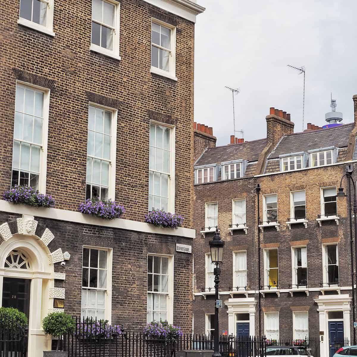 A street view of classic London townhouses with brown brick facades, white-trimmed sash windows, and purple flowers in window boxes, under a cloudy sky.