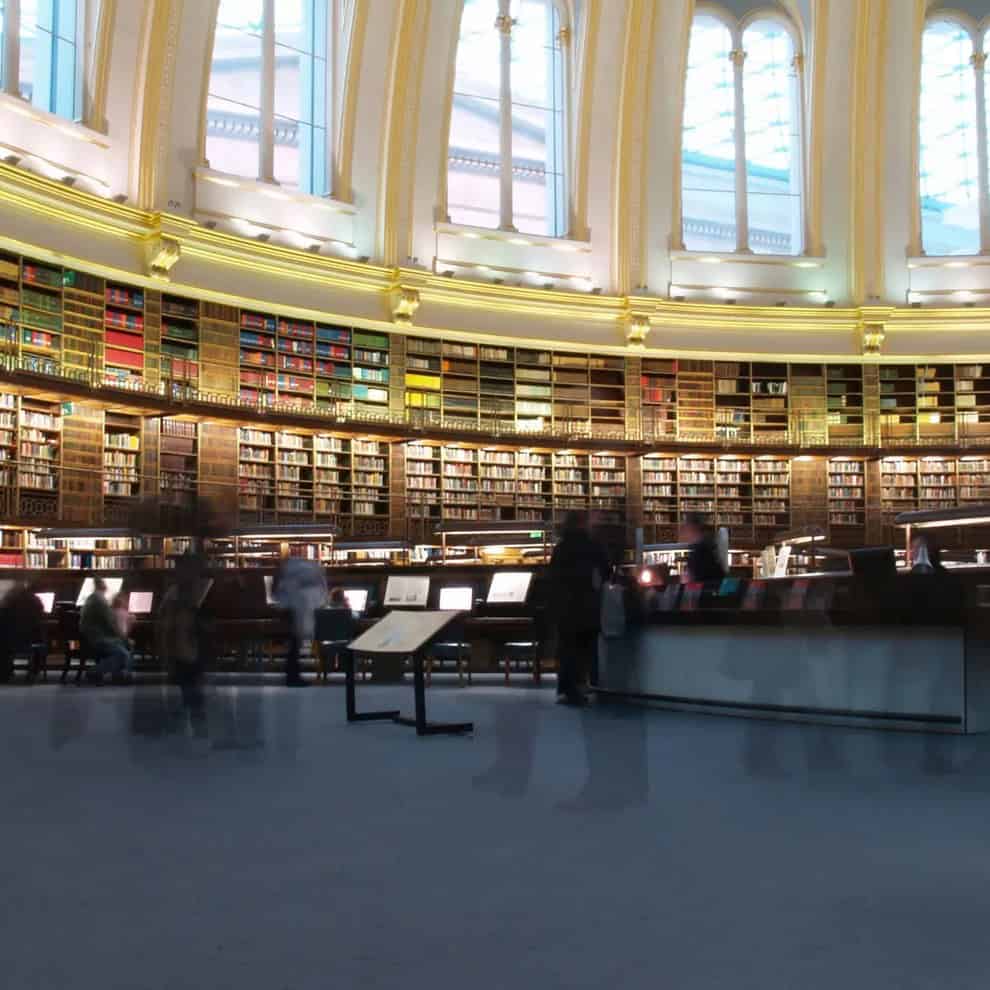 Large, circular library with high arched windows and rows of bookshelves lining the walls. Several people are browsing or working at desks, with some motion blur suggesting movement. Soft, ambient lighting fills the space.