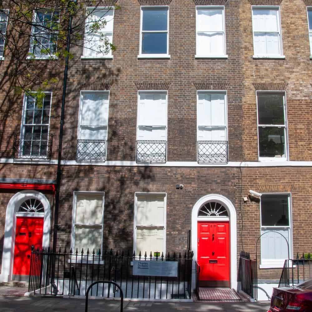 A row of brick townhouses with white-trimmed windows and two bright red doors; the windows have white shutters, and black iron railings line the sidewalk in sunlight with tree shadows.