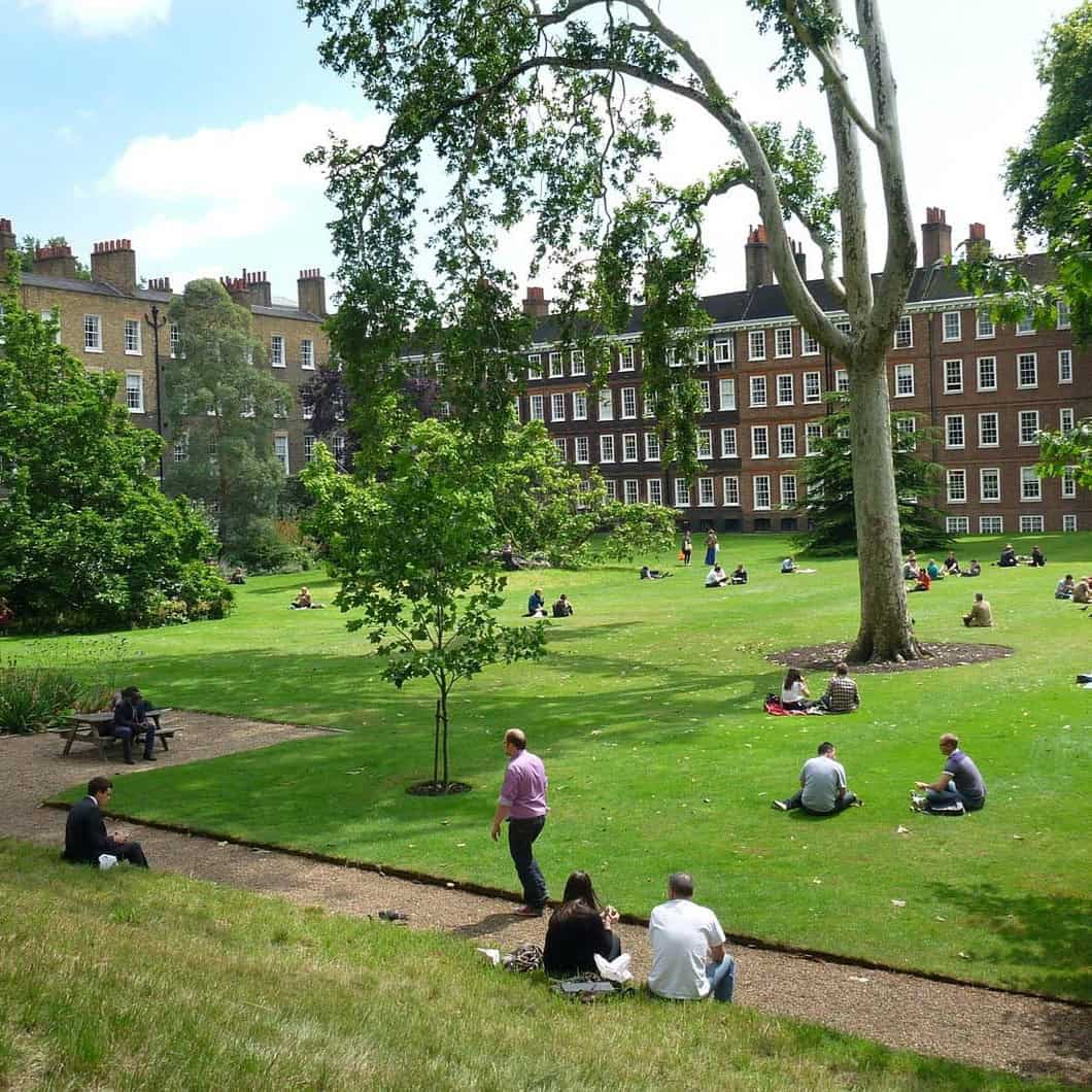 People sit and relax on a large, green lawn surrounded by historic brick buildings and tall trees on a sunny day. Some walk along a gravel path, while others sit in small groups or alone on the grass.