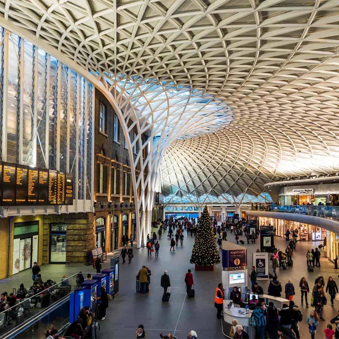 King’s Cross railway station interior with a vaulted, lattice-patterned ceiling, large Christmas tree in the center, people walking or waiting, electronic departure boards, and shops lining the walls.