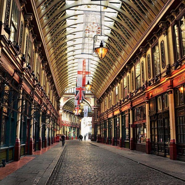 A covered, historic shopping arcade with arched glass ceilings, ornate architecture, hanging lanterns, and British flags. Cobblestone street with shop fronts on both sides. Few people are visible in the distance.