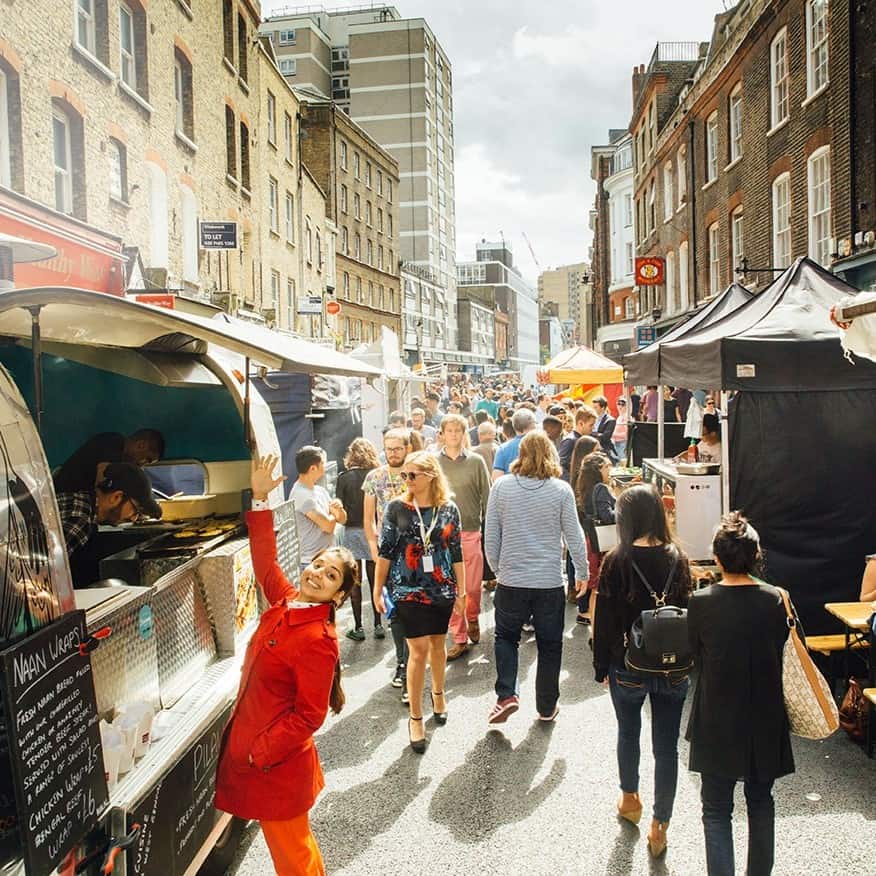 A busy outdoor street market with many people walking, food stalls on both sides, and a smiling woman in a red coat waving at the camera. The scene is lively and set on a sunny day in an urban area.