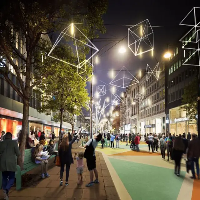 A lively city street at night with people walking and sitting. Trees line the sidewalks, and geometric light sculptures hang above, illuminating the scene. Buildings with lit windows and shops create a vibrant urban atmosphere.