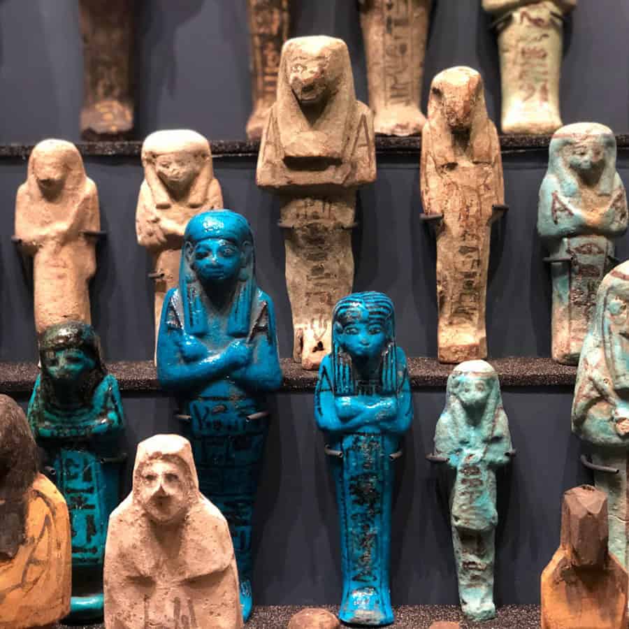 A display of ancient Egyptian shabti figurines, mostly brown and beige, with two prominent blue figures in the center, all standing upright against a dark background.