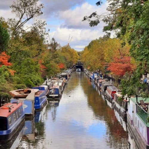 A narrow canal lined with colorful houseboats on both sides, surrounded by autumn trees with orange, yellow, and green leaves under a partly cloudy sky.