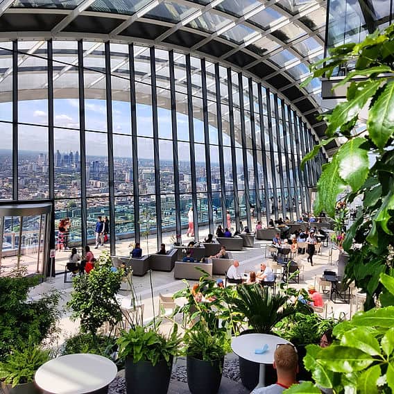 People relax and enjoy views of London through large glass windows inside a spacious, modern, indoor garden with seating areas and lush green plants, under an arched glass roof.