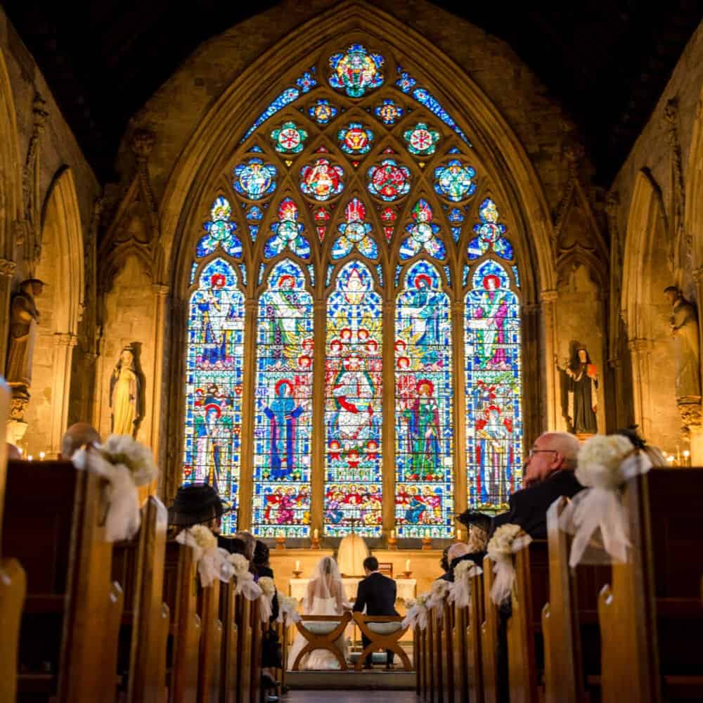 A couple sits at the altar in a church with high ceilings, surrounded by guests. The backdrop is a large, vibrant stained glass window, and the pews are decorated with white flowers.