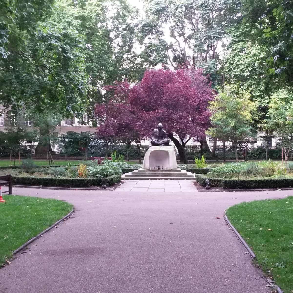 A statue on a pedestal stands in the center of a park surrounded by trees, shrubs, and a pathway, with a vibrant purple-leaved tree behind the statue.