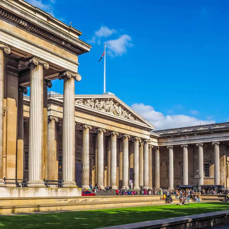 The British Museum’s grand neoclassical facade with tall columns, intricate pediment, and a flagpole, under a bright blue sky. People are gathered by the entrance and walking across the green lawn.