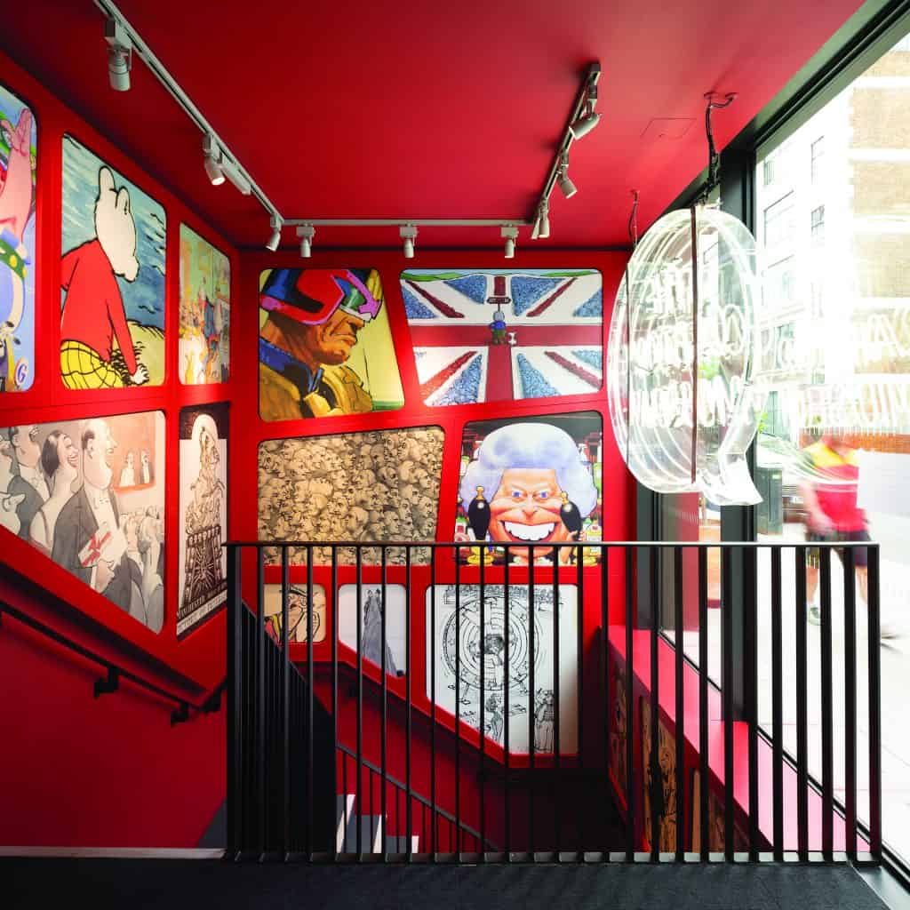 A brightly lit stairwell with red walls decorated with vibrant comic art and British-themed illustrations, including a Union Jack. A large speech bubble-shaped light hangs by the glass window overlooking the street.