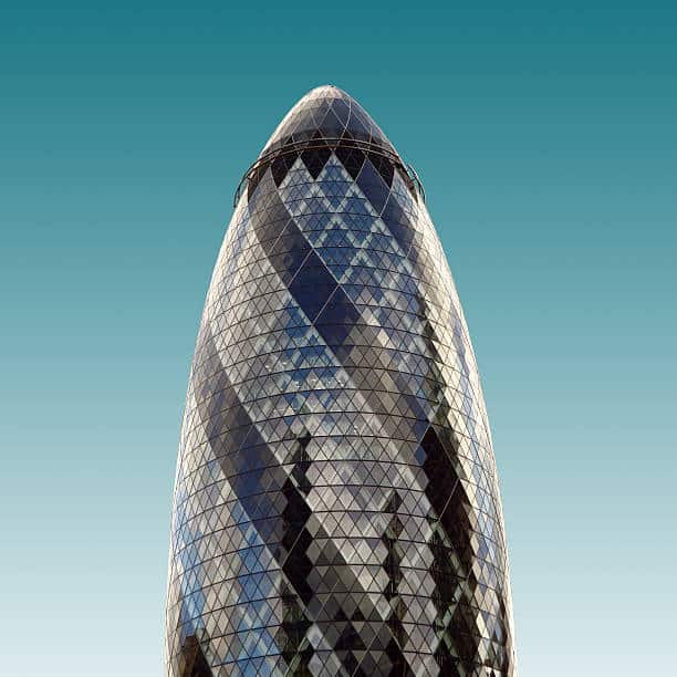 A close-up view of the Gherkin building in London, featuring its distinctive curved shape and diamond-patterned glass façade, set against a clear blue sky.