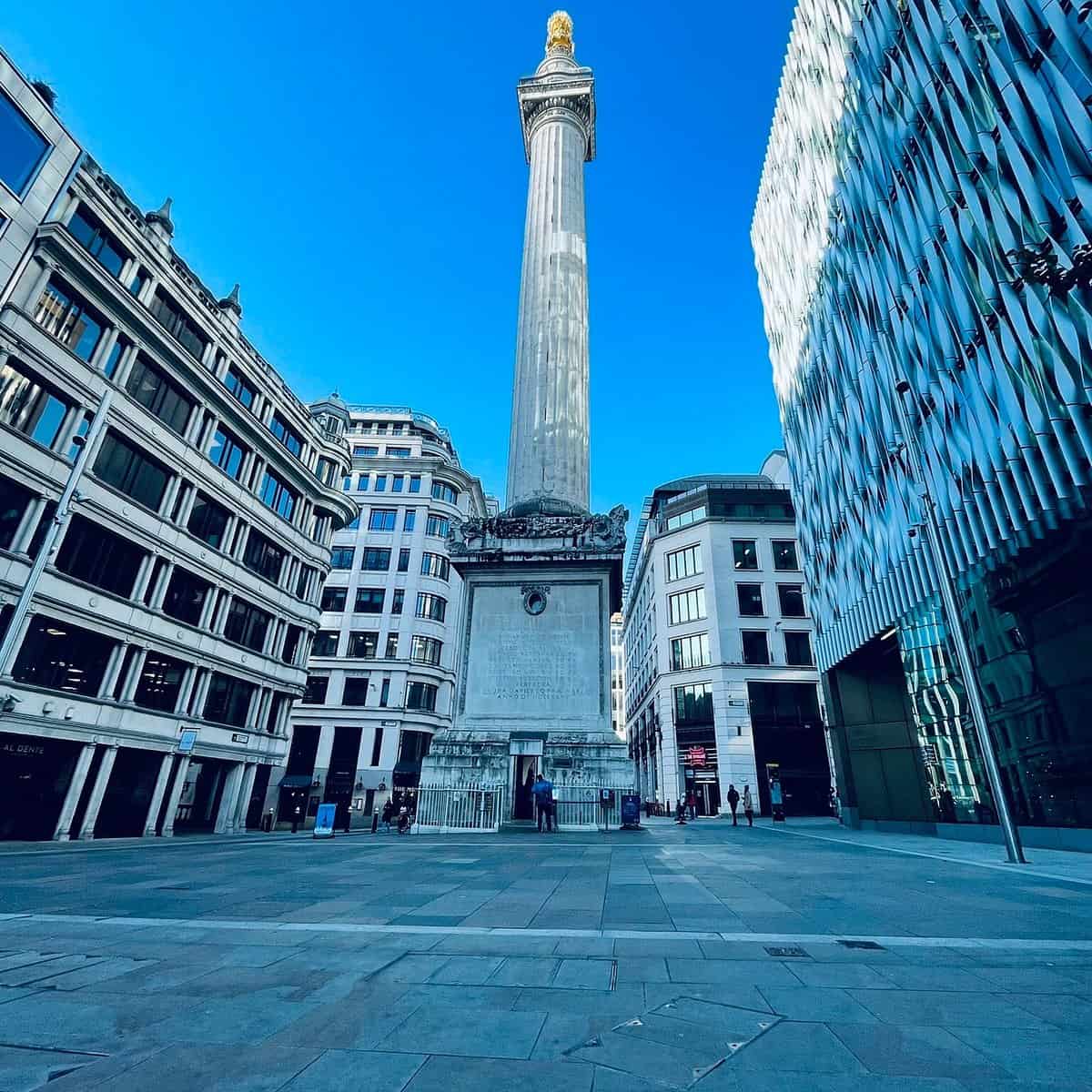 A tall stone column with a golden top stands in an open square surrounded by modern and historic buildings under a clear blue sky. People are visible near the base of the monument.