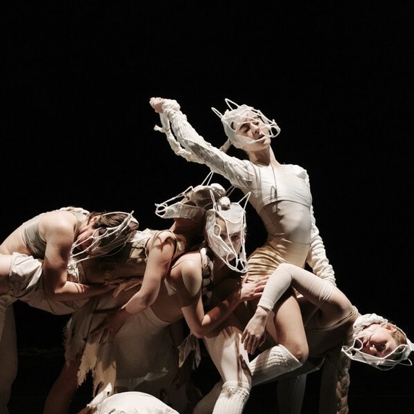 Five performers in abstract white costumes and headpieces pose dramatically on stage, forming an intricate, sculptural arrangement against a dark background. Their movements and attire evoke a sense of avant-garde performance art.
