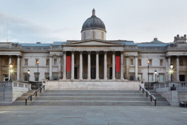 The image shows the front view of the National Gallery in London, featuring neoclassical architecture with tall columns, a central dome, and two red banners hanging between the columns. The square in front is empty.