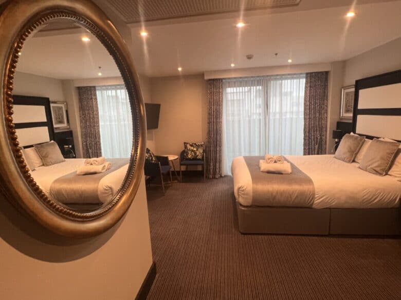 A hotel room with two double beds, neutral bedding, a round mirror on the wall, large windows with curtains, a small table with chairs, and folded towels placed on each bed.