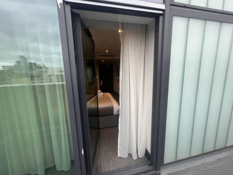 A view into a modern hotel room through an open glass door, with white curtains partly drawn. Inside, a neatly made bed with folded towels is visible, and city buildings can be seen through the glass on the left.