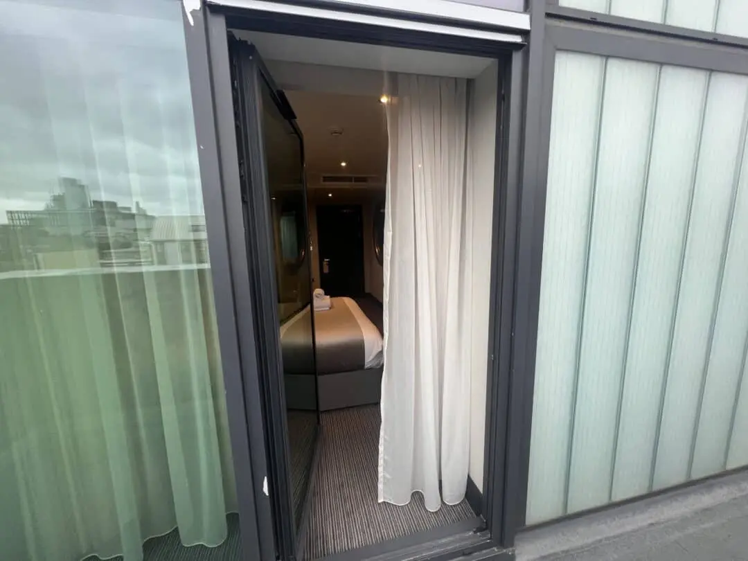 A view into a modern hotel room through an open glass door, with white curtains partly drawn. Inside, a neatly made bed with folded towels is visible, and city buildings can be seen through the glass on the left.