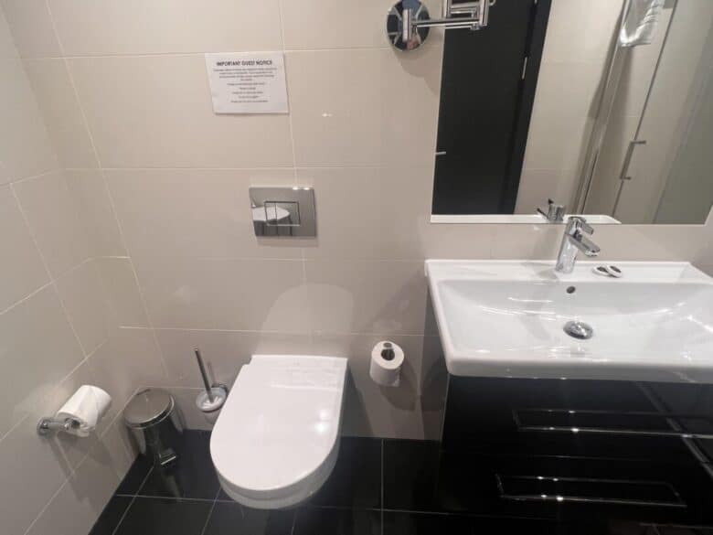 Modern bathroom with a wall-mounted toilet, toilet brush, trash can, toilet paper holders, and a white sink with a mirror above it. The walls are tiled white and the floor is black. A sign is posted above the toilet.