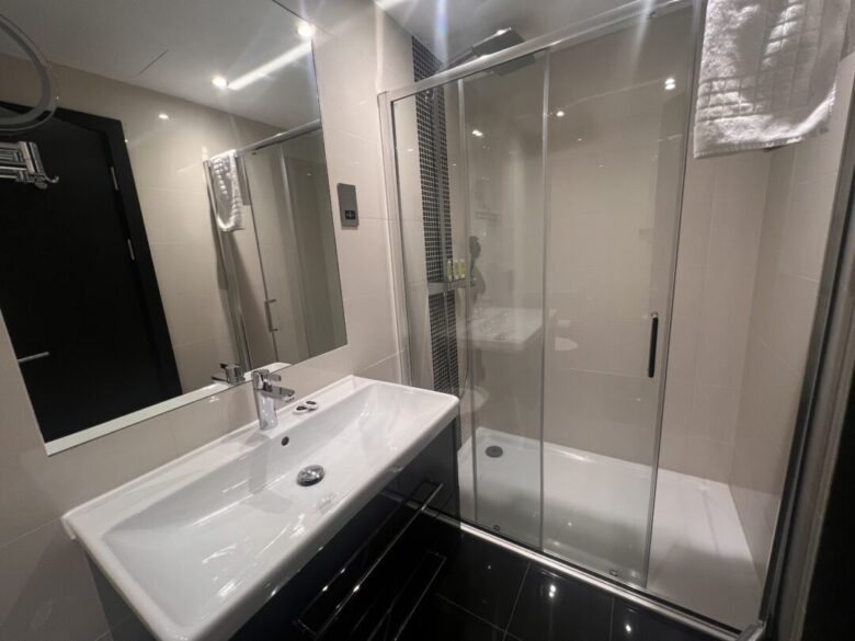 Modern bathroom with a large mirror, white sink and vanity, and a glass-enclosed shower. A towel hangs near the shower, and the room has light-colored walls and dark floor tiles.