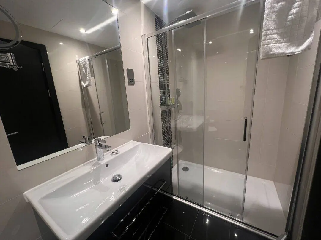 Modern bathroom with a large mirror, white sink and vanity, and a glass-enclosed shower. A towel hangs near the shower, and the room has light-colored walls and dark floor tiles.