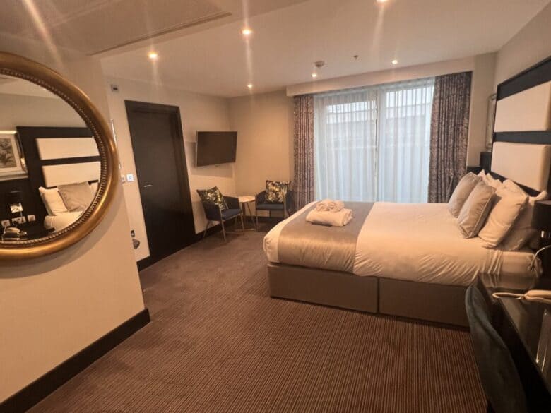 A cozy hotel room with a double bed, two chairs and a small table, a TV mounted on the wall, large window with curtains, and a round mirror on the wall reflecting part of the room.