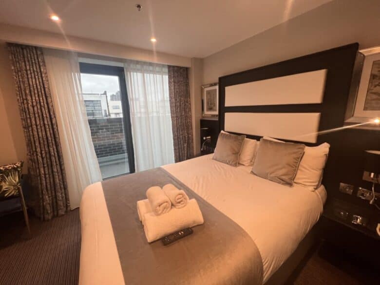 A modern hotel room with a double bed, three gray cushions, folded towels, and a TV remote on the bed. Floor-to-ceiling curtains cover a glass door leading to a balcony with a city view.