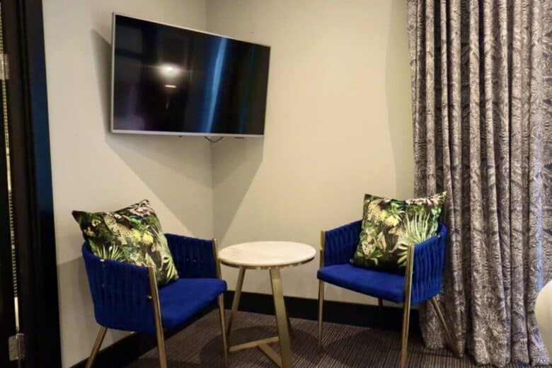 A small seating area with two blue chairs featuring tropical-print pillows, a round marble-top table between them, and a wall-mounted flat-screen TV above, next to patterned curtains.