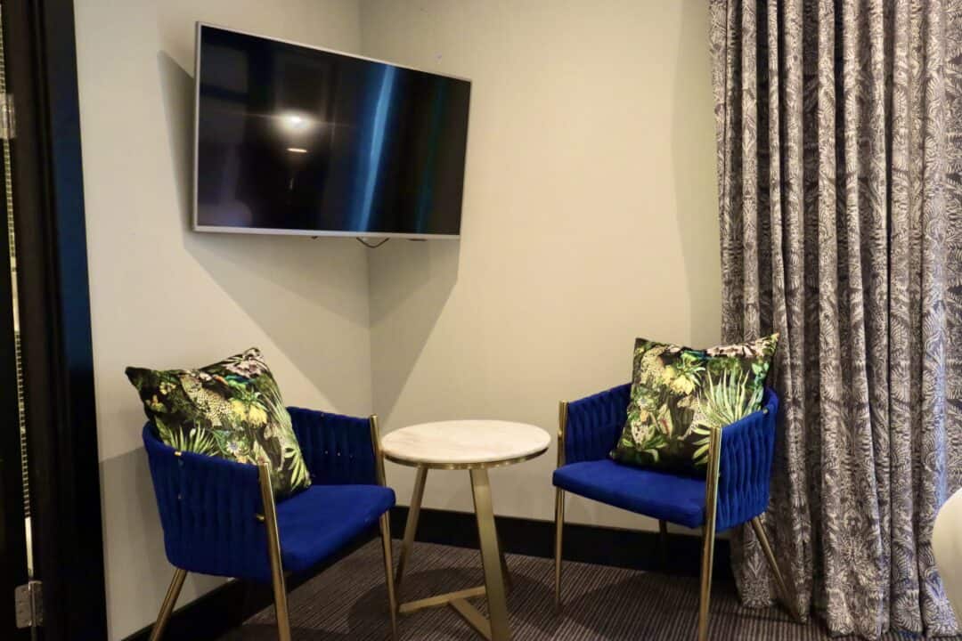 A small seating area with two blue chairs featuring tropical-print pillows, a round marble-top table between them, and a wall-mounted flat-screen TV above, next to patterned curtains.