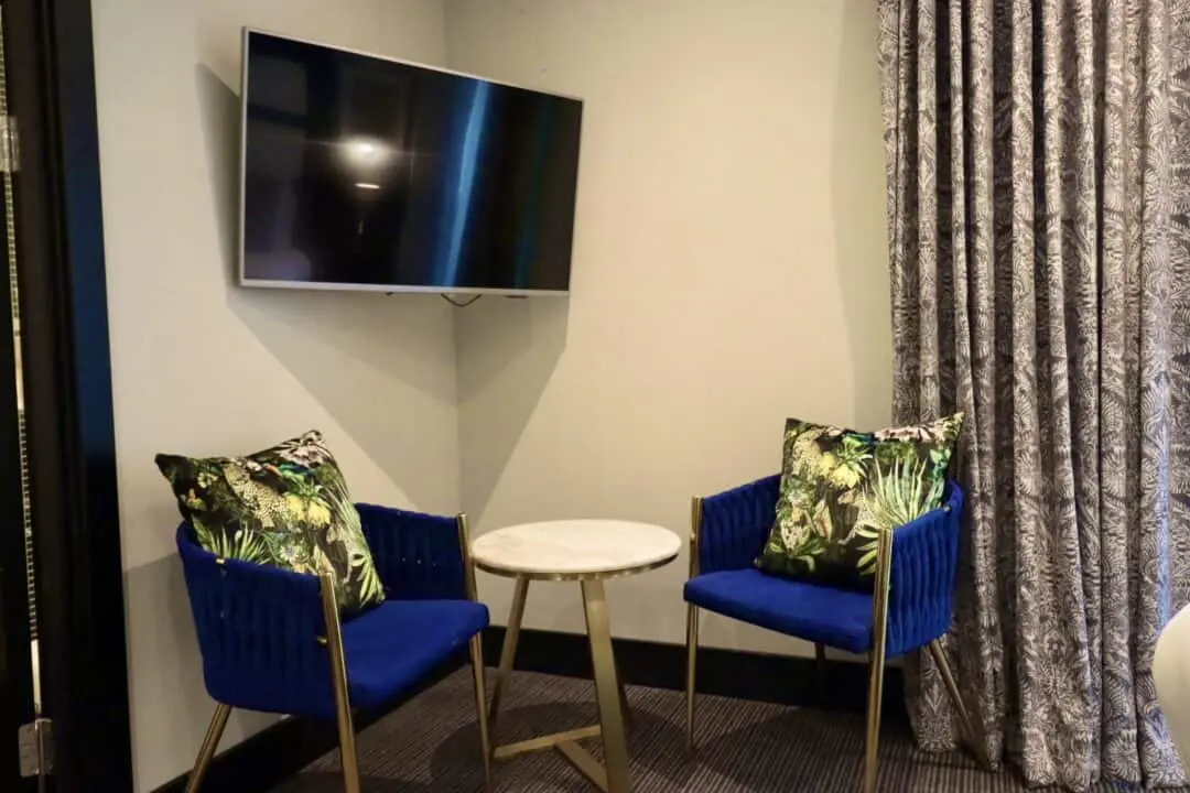 A small seating area with two blue chairs featuring tropical-print pillows, a round marble-top table between them, and a wall-mounted flat-screen TV above, next to patterned curtains.