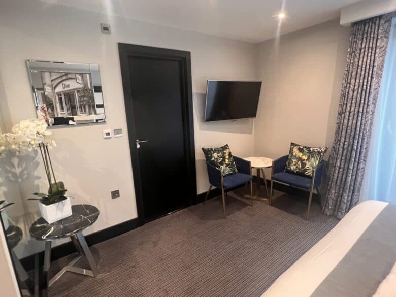 Modern hotel room with gray walls, a black door, a wall-mounted TV, two blue chairs with floral cushions around a small round table, a potted orchid, a patterned curtain, and a decorative mirror.