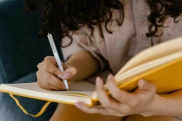 A person with curly hair writes in a yellow notebook, jotting down 5 Productivity Hacks While Staying at a Hotel. Seated and wearing a light-colored, long-sleeve shirt, they hold the open notebook in one hand and write with a white pen in the other.