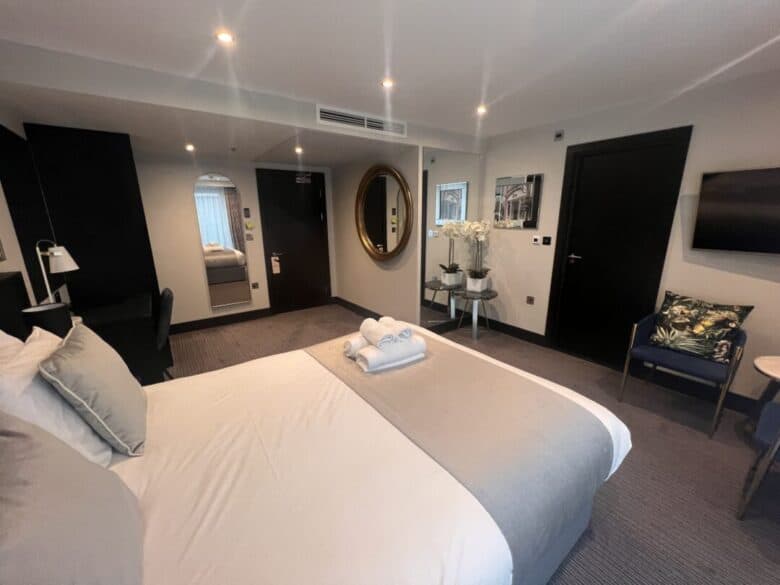A modern hotel room with a neatly made double bed, folded towels on top, a desk, wall-mounted TV, round mirror, and decorative plants. The room features neutral tones and soft lighting.