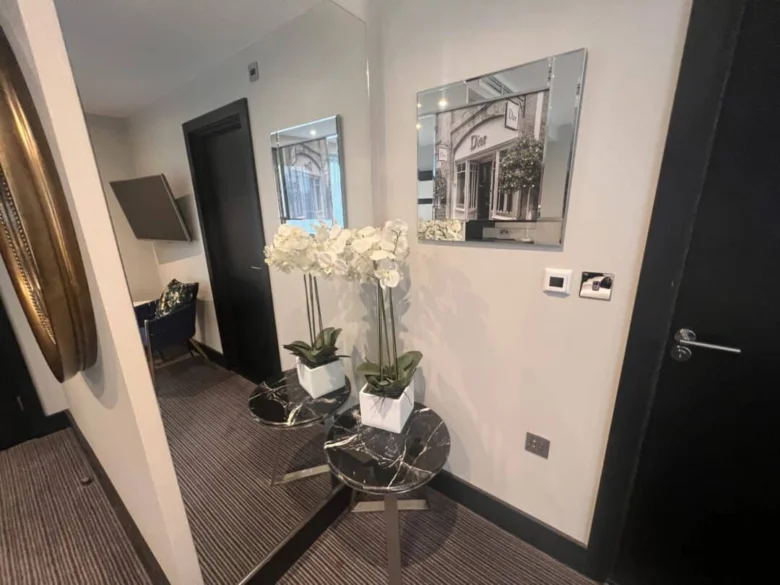 A hallway with a black marble table holding white orchids in a square pot, placed in front of a large mirror. A framed photo, light switch, and door are visible on the light-colored wall.