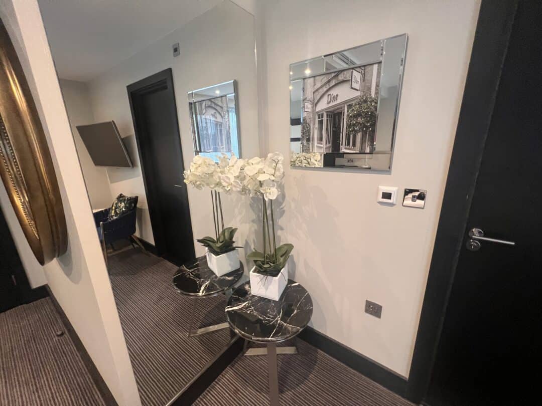 A hallway with a black marble table holding white orchids in a square pot, placed in front of a large mirror. A framed photo, light switch, and door are visible on the light-colored wall.