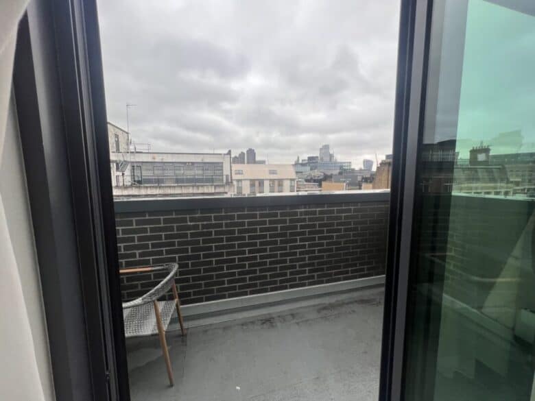 View through a glass door onto a small balcony with a gray brick wall, a chair, and a cityscape of modern and older buildings under a cloudy sky in the background.