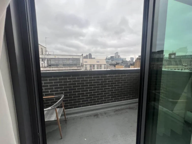 View through a glass door onto a small balcony with a gray brick wall, a chair, and a cityscape of modern and older buildings under a cloudy sky in the background.