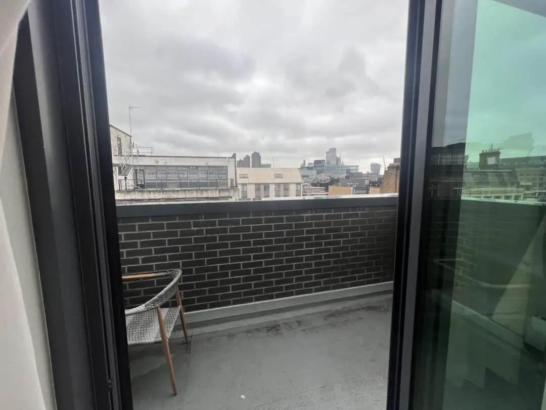View through a glass door onto a small balcony with a gray brick wall, a chair, and a cityscape of modern and older buildings under a cloudy sky in the background.