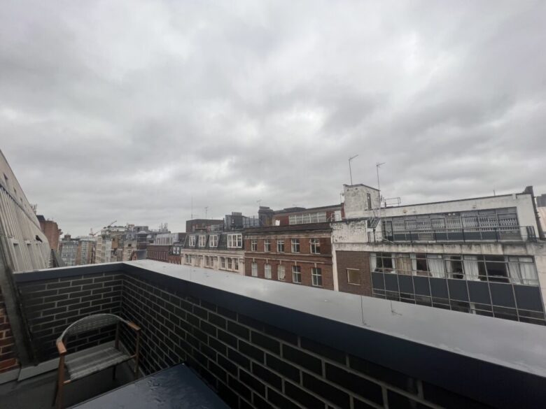 View from a balcony on a cloudy day, showing brick buildings with multiple windows. The balcony has a brick railing and a chair. The sky is overcast with grey clouds.