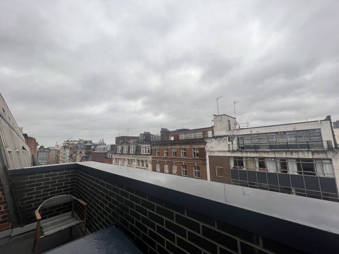 View from a balcony on a cloudy day, showing brick buildings with multiple windows. The balcony has a brick railing and a chair. The sky is overcast with grey clouds.