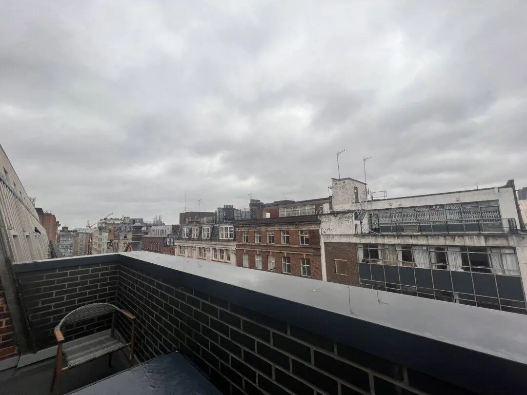 View from a balcony on a cloudy day, showing brick buildings with multiple windows. The balcony has a brick railing and a chair. The sky is overcast with grey clouds.