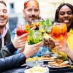 Five friends sit outdoors at a table, smiling and raising colorful cocktails in a toast. Plates of food, including chips and sandwiches, are in front of them. The atmosphere is cheerful and lively.