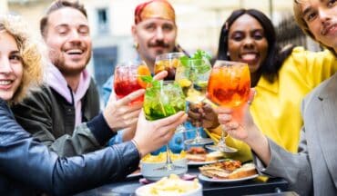 Five friends sit outdoors at a table, smiling and raising colorful cocktails in a toast. Plates of food, including chips and sandwiches, are in front of them. The atmosphere is cheerful and lively.