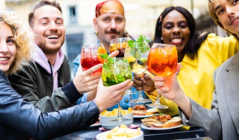 Five friends sit outdoors at a table, smiling and raising colorful cocktails in a toast. Plates of food, including chips and sandwiches, are in front of them. The atmosphere is cheerful and lively.