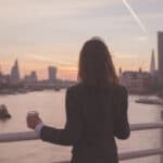 A person in a dark suit stands on a bridge overlooking a river at sunset, holding a coffee cup, with city buildings and a jet trail in the sky in the background.