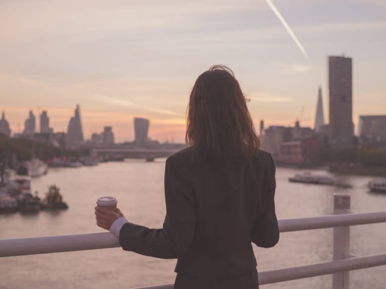 A person in a dark suit stands on a bridge overlooking a river at sunset, holding a coffee cup, with city buildings and a jet trail in the sky in the background.