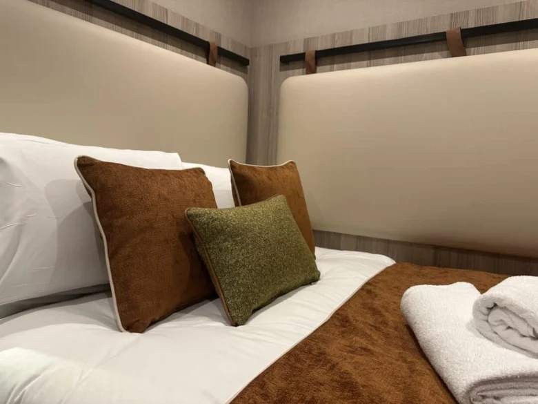 A neatly made bed with white sheets, brown and green accent pillows, a brown blanket, and folded white towels, set against a padded beige headboard and wooden wall panels.
