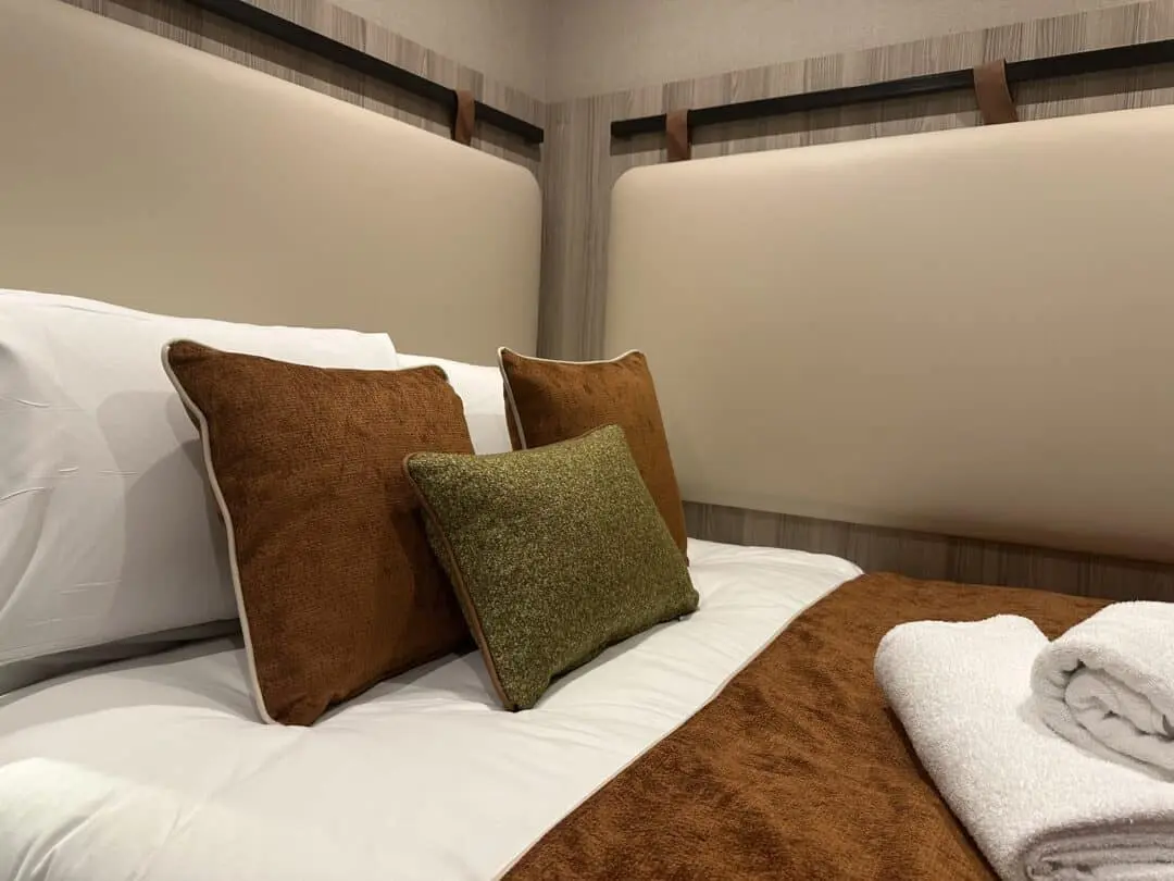 A neatly made bed with white sheets, brown and green accent pillows, a brown blanket, and folded white towels, set against a padded beige headboard and wooden wall panels.